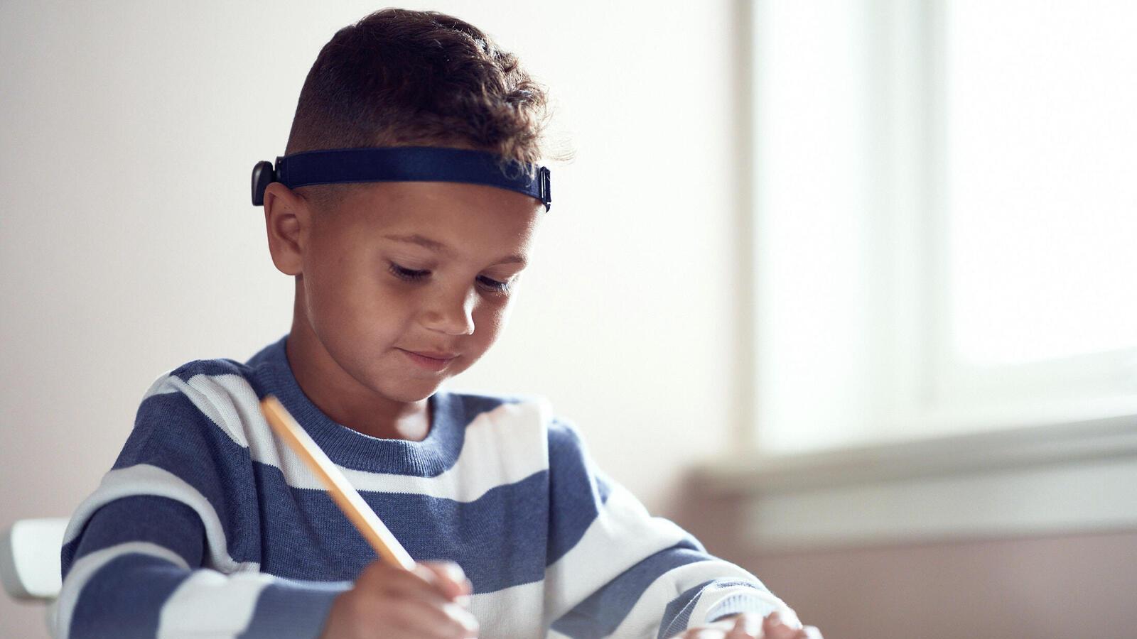 Young boy wearing a bone conduction device on a headband sitting drawing or writing
