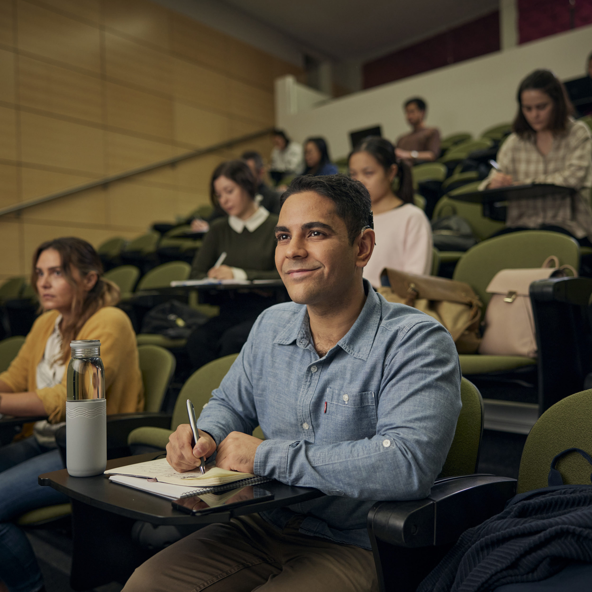 Male student sitting in lecture hall with other students writing in a notebook